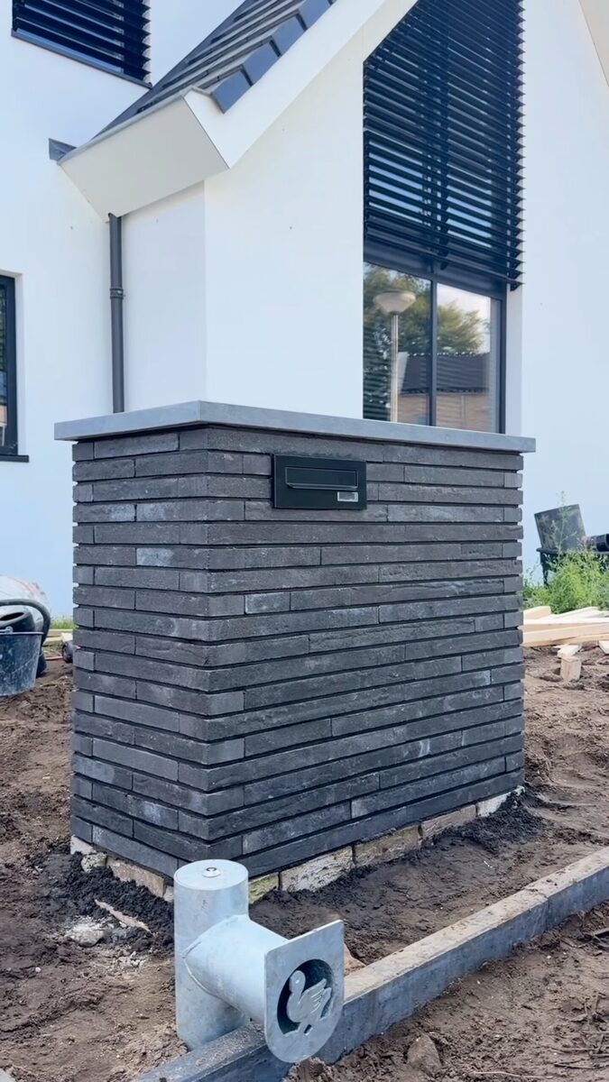 Gemetselde brievenbuszuil van donkergrijze langformaat stenen met een ingemetselde brievenbus, voor een witte nieuwbouwwoning